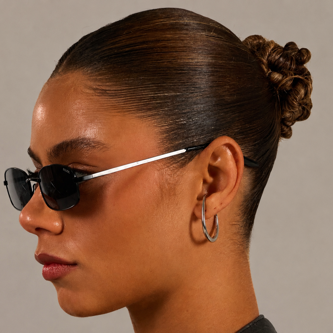 A close-up side profile of a model wearing black cat-eye sunglasses with thin silver temples and a high bun hairstyle, set against a solid neutral gray background.