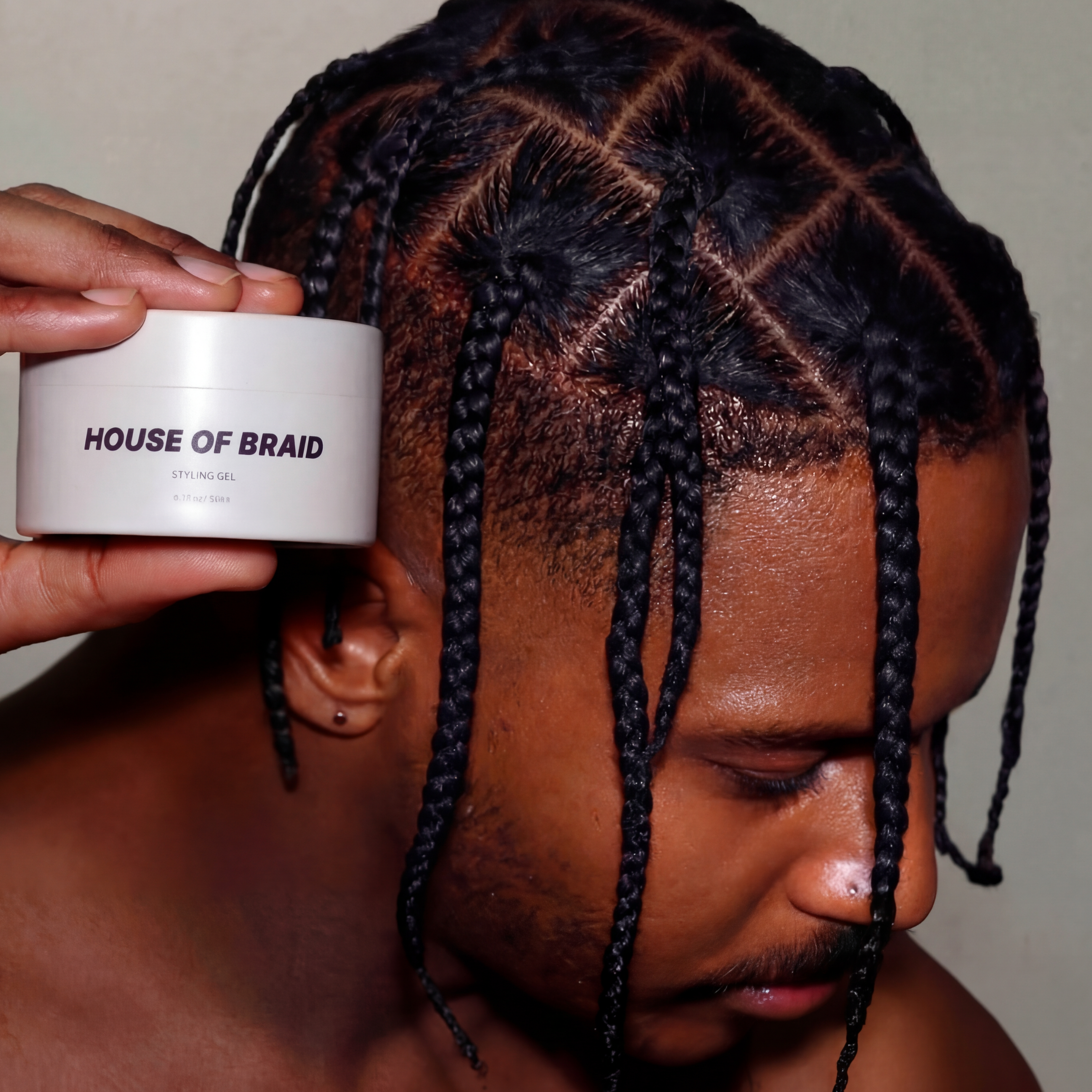 A close-up of a man with intricate black braids and a geometric grid pattern on his scalp, holding a white jar of House of Braid hair product near his head.