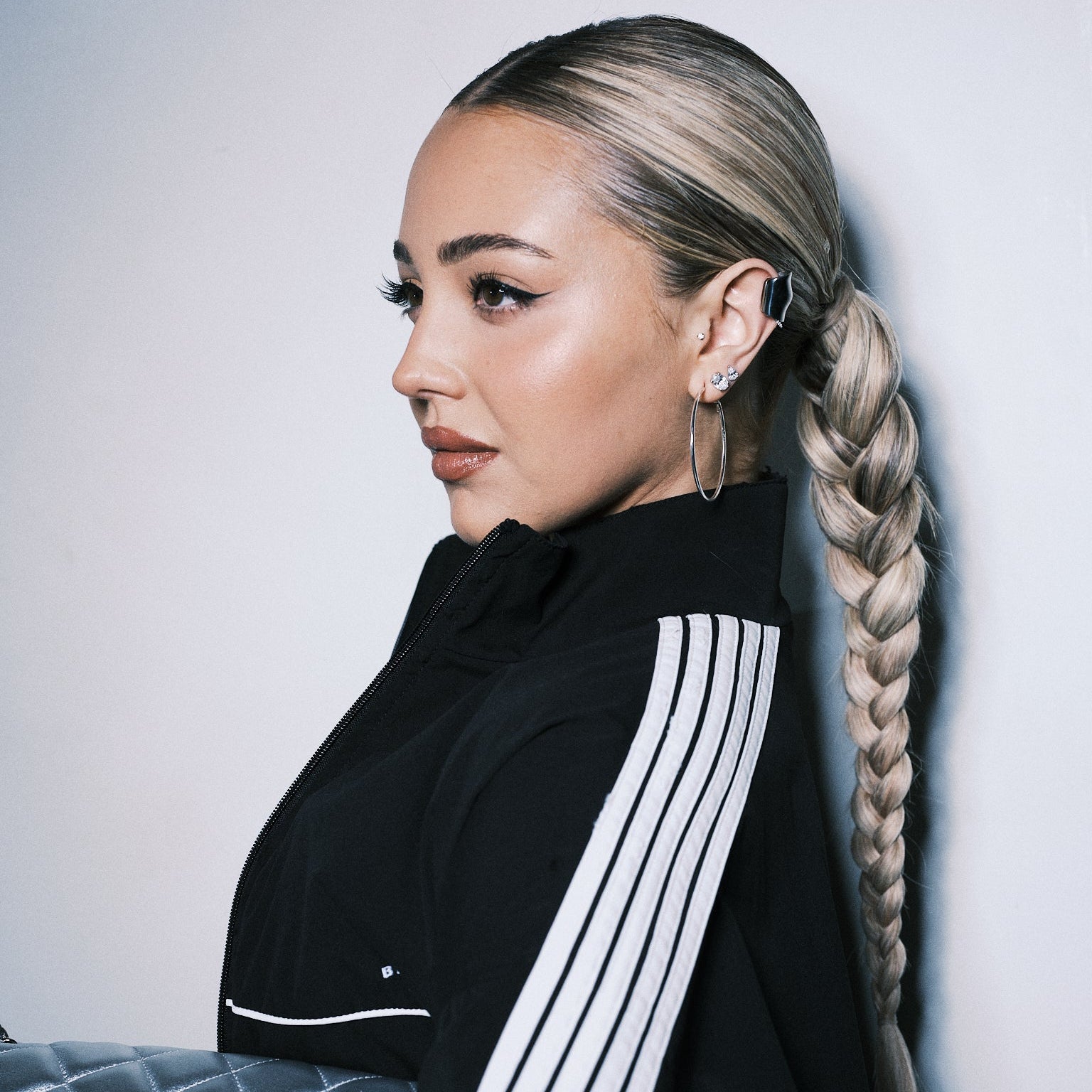 A woman with blonde-highlighted hair in a long braid, wearing a black mock-neck top with white vertical stripes on the sleeves, posed next to a silver quilted handbag against a light gray wall.